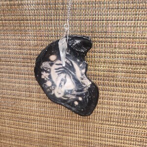 May include: A unique pendant necklace featuring a black, shell-shaped charm with a white and gray design. A clear crystal hangs from a silver chain, adding a touch of elegance. The charm has a detailed, artistic design.