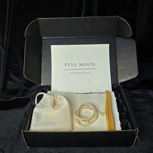 Puede incluir: Una caja negra que contiene un kit de ritual de luna llena. El kit incluye una pequeña bolsa con cordón, una cuerda enrollada, un palo de madera y una tarjeta con el texto "FULL MOON A Release Ritual". Los artículos están dispuestos sobre una tela blanca.