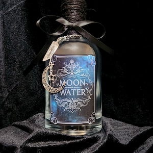 Può includere: Una bottiglia di vetro trasparente con l'etichetta "MOON WATER" con un design celeste. La bottiglia è adornata con un nastro nero, spago e un ciondolo d'argento con una luna crescente e un pendente di cristallo. La bottiglia ha un tappo nero.