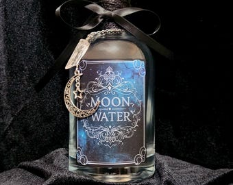 Puede incluir: Una botella de vidrio transparente con la etiqueta "MOON WATER" con un diseño celestial. La botella está adornada con una cinta negra, hilo y un dije plateado con una luna creciente y un colgante de cristal. La botella tiene una tapa negra.