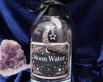 Puede incluir: Una botella de vidrio transparente con una etiqueta negra que dice "Moon Water". La botella está decorada con una cinta negra y un dije de plata en forma de media luna con una estrella.