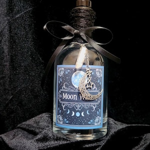 Puede incluir: Una botella de vidrio transparente con una tapa negra y una cinta negra atada alrededor del cuello. La botella tiene una etiqueta con un diseño de luna y estrellas y las palabras "Moon Water". Un dije plateado con una luna creciente y un cristal cuelga de la botella.