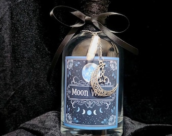 Puede incluir: Una botella de vidrio transparente con una tapa negra y una cinta negra atada alrededor del cuello. La botella tiene una etiqueta con un diseño de luna y estrellas y las palabras "Moon Water". Un dije plateado con una luna creciente y un cristal cuelga de la botella.