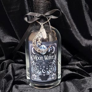 May include: A clear glass bottle with a black label that reads "Moon Water". The bottle is adorned with a silver crescent moon charm and a crystal pendant. The bottle is tied with a black ribbon.