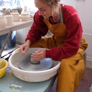 Może przedstawiać: Osoba w czerwonym swetrze i żółtym fartuchu używa koła garncarskiego do formowania kawałka gliny. Koło jest białe i ma szary podnóżek. Osoba ma na sobie niebieskie dżinsy i czarne buty sportowe.