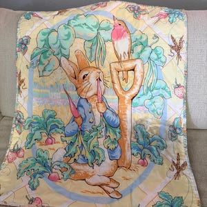 May include: A yellow, square baby blanket featuring a whimsical illustration of a rabbit in a blue jacket eating a carrot, with a bird perched on a shovel. The blanket has a border with strawberries, wheat, and other garden elements.