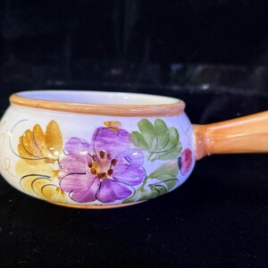 May include: A white ceramic soup bowl with a handle, adorned with hand-painted floral designs in purple, yellow, and green. The rim and handle are painted in shades of orange. A decorative kitchenware item.