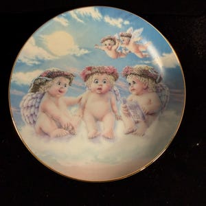 May include: Decorative plate featuring cherubic figures with wings, floral wreaths, and a blue sky background. The plate has a gold rim and depicts cherubs on clouds, with one holding a book titled "How to Fly".