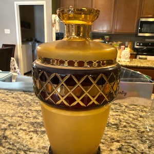 May include: A vintage amber glass decanter with a decorative stopper. The decanter features a geometric diamond pattern in a dark red color, with gold accents and a frosted glass base. The overall design is ornate and elegant.