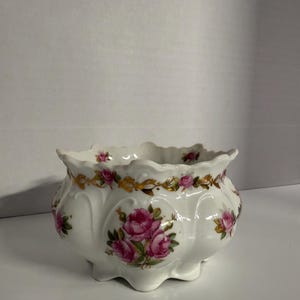 May include: White porcelain planter with a scalloped rim and a floral design. The planter features pink roses and gold accents. The planter has a rounded shape and sits on three small feet.