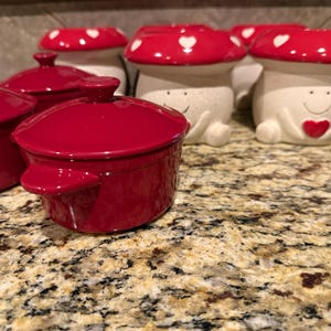 May include: A collection of ceramic mushroom-shaped containers and small red lidded pots. The mushroom tops are red with white heart accents. The mushroom bodies are off-white with smiling faces and heart details. The pots are a glossy red.