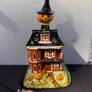 May include: A ceramic Halloween house figurine featuring a black roof with white dots, orange walls, and a jack-o'-lantern topper wearing a black witch hat. The base is green with multiple orange pumpkins and a sign reading Pumpkin Patch Palace.