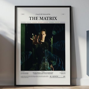 May include: A framed poster for the film "The Matrix" with a black frame. The poster shows a person reaching out, with the film's title at the top. The poster is displayed against a white wall, with furniture and plants in the background.