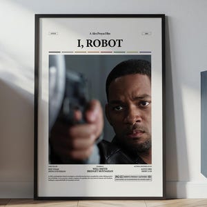 Puede incluir: Un póster enmarcado de la película "I, Robot" con Will Smith. El póster tiene un marco negro y está sobre un suelo de madera. El póster está contra una pared blanca, con un mueble y plantas a ambos lados. El texto del póster es visible.