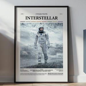 Pode incluir: Pôster emoldurado do filme "Interstellar" com um astronauta em frente a uma paisagem montanhosa nevada. O pôster tem uma moldura preta e é exibido em uma sala com móveis de madeira.