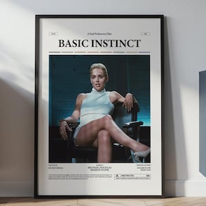 May include: Framed movie poster for "Basic Instinct" featuring a woman in a light blue outfit. The poster has a black frame and is displayed on a wall. The poster is in a room with a wooden floor and furniture.
