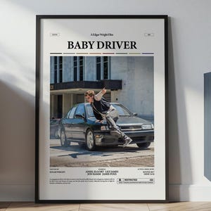 May include: A framed movie poster for "Baby Driver" featuring a scene with a car and a person. The poster has a black frame and is displayed against a white wall. The poster's design includes the movie title and a photograph.