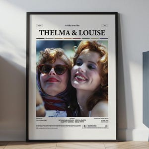 Puede incluir: Un póster enmarcado de la película "Thelma & Louise" con dos mujeres. Una lleva gafas de sol y un pañuelo, la otra sonríe. El póster está en un marco negro, apoyado en una pared blanca, con un mueble claro y un mueble azul oscuro en el fondo.