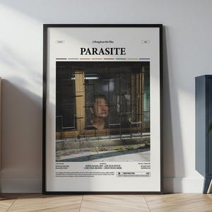 Pode incluir: Um cartaz emoldurado do filme "Parasite" com uma moldura preta, encostado a uma parede branca. O cartaz apresenta uma pessoa olhando por uma janela. O título "PARASITE" está no topo. O cartaz está numa sala com mobiliário.