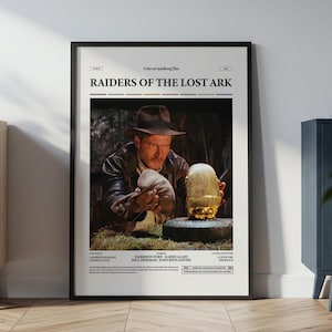 May include: Framed movie poster for "Raiders of the Lost Ark." The poster features a scene with a man in a fedora examining a golden artifact. The poster is in a black frame and is displayed on a wall.