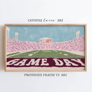 Puede incluir: Una impresión enmarcada de un estadio de fútbol con un campo verde y asientos rosas. Las palabras "GAME DAY" se muestran en blanco sobre un fondo granate. También se ve el texto "gemma loves art" y "PREMIUM FRAME TV art".