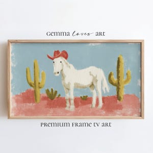 Puede incluir: Una impresión artística enmarcada que presenta un caballo blanco con un sombrero de vaquero rojo, entre dos cactus amarillos. El fondo es un cielo azul claro y un paisaje desértico rojo. El texto "gemma loves art" y "PREMIUM FRAME TV art" son visibles.