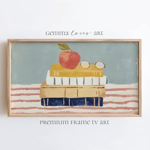 May include: A framed print featuring a stack of books, a red apple, and round eyeglasses. The books are in shades of yellow, white, and blue. The background is a light blue with a red and white striped pattern at the bottom. The text "gemma loves art" and "PREMIUM FRAME TV art" are visible.