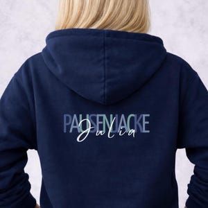 May include: Navy blue hoodie with the text "PAUSENJACKE Julia" in blue and white lettering on the back. The word "NAVY" is in a box in the upper left corner. The hoodie has a hood and long sleeves.