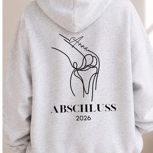 May include: A light gray heathered hoodie featuring a black line art illustration of a knee joint on the back. The design includes the name Anna written in script above the illustration, with the text ABSCHLUSS 2026 printed in black below it.