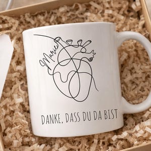 May include: White ceramic mug with a black line drawing of a heart and the name "Marie". The mug also has the text "DANKE, DASS DU DA BIST". The mug is in a cardboard gift box with shredded paper filler, alongside a tag with a heart and cross symbol.