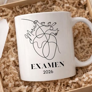 May include: White ceramic mug with a black line drawing of a heart and the word "EXAMEN" and the year "2026". The mug is in a gift box with shredded paper. A tag with a heart and cross is attached.