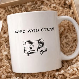 May include: A white ceramic mug featuring the black text "wee woo crew" above a line art illustration of an ambulance with a cross symbol. The mug is nestled inside a square cardboard gift box filled with shredded tan paper filler material.