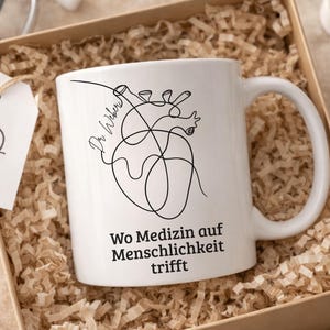 May include: White ceramic mug with a black line drawing of a heart and the words "Wo Medizin auf Menschlichkeit trifft". The mug is in a gift box with shredded paper. A tag with a heart and cross is attached.