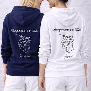 May include: Two hoodies, one navy blue and one white, with the text "Pflegeexamen 2026" and a line art heart design on the back. The navy hoodie has the name "Marie" below the design, and the white hoodie has the name "Anna".