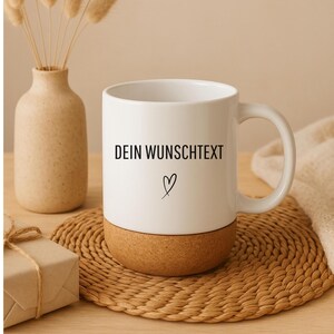 May include: White ceramic mug with a cork base, featuring the text "DEIN WUNSCHTEXT" and a heart symbol. The mug sits on a woven coaster, with a vase of dried flowers and a wrapped gift in the background.