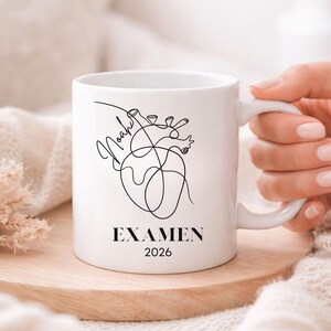 May include: White ceramic mug featuring a black line art illustration of a heart with the name "Noah" and the word "EXAMEN" with the year "2026". The mug is on a wooden coaster.