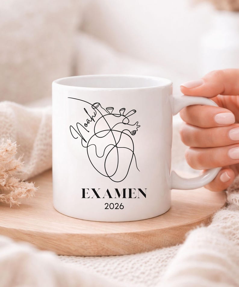 May include: White ceramic mug featuring a black line art illustration of a heart with the name "Noah" and the word "EXAMEN" with the year "2026". The mug is on a wooden coaster.