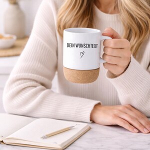 May include: White ceramic mug with a cork base, featuring the text "DEIN WUNSCHTEXT" and a heart symbol. The mug has a handle and is held by a person. A notebook and pen are in the foreground.