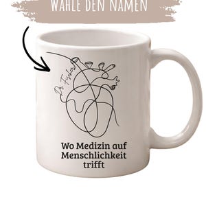 May include: White ceramic mug with a black line art illustration of a heart. The mug has the text "Wo Medizin auf Menschlichkeit trifft" and "Dr. Fischer" with the words "W&auml;hle den Namen" above the mug.