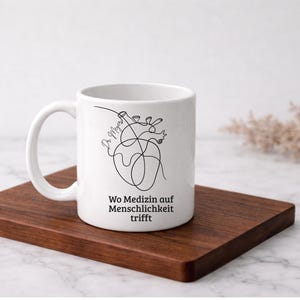 May include: White ceramic mug with a black line drawing of a heart and the text "Wo Medizin auf Menschlichkeit trifft". The mug is on a wooden tray.