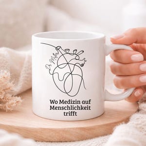 May include: White ceramic mug with a black line drawing of a heart and the text "Wo Medizin auf Menschlichkeit trifft". The mug is on a wooden coaster.