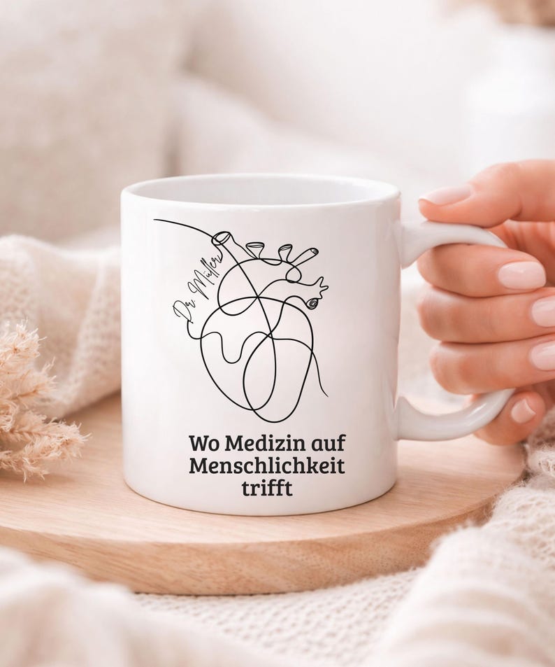 May include: White ceramic mug with a black line drawing of a heart and the text "Wo Medizin auf Menschlichkeit trifft". The mug is on a wooden coaster.