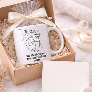 May include: A white ceramic mug with a black line drawing of a heart and the text "Wo Medizin auf Menschlichkeit trifft" inside a gift box. The box is filled with shredded paper and tied with a cream-colored ribbon.