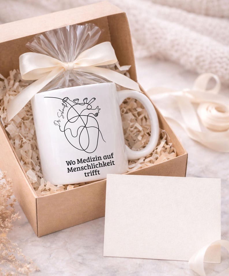 May include: A white ceramic mug with a black line drawing of a heart and the text "Wo Medizin auf Menschlichkeit trifft" inside a gift box. The box is filled with shredded paper and tied with a cream-colored ribbon.