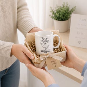 May include: A white ceramic mug with a black line drawing of a heart and the text "Wo Medizin auf Menschlichkeit trifft" is presented in a gift box. A small tag with a heart and the words "Danke f&uuml;r alles" is attached.