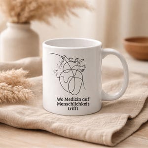 May include: White ceramic mug with a black line drawing of a heart and the words "Wo Medizin auf Menschlichkeit trifft". The mug has a curved handle and is sitting on a beige cloth.