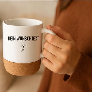 May include: White ceramic mug with a cork base, featuring the text "DEIN WUNSCHTEXT" and a heart symbol. The mug has a curved handle and is held by a person wearing a brown sweater.