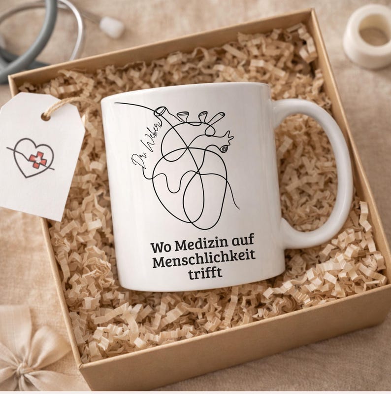 May include: White ceramic mug with a black line drawing of a heart and the words "Wo Medizin auf Menschlichkeit trifft". The mug is in a gift box with shredded paper. A tag with a heart and cross is attached.