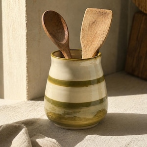 May include: A ceramic utensil holder with a cream-colored base and horizontal olive green stripes, containing one wooden spoon and one wooden spatula. The container has a rounded, tapered shape with a smooth, glossy finish and a wide opening.