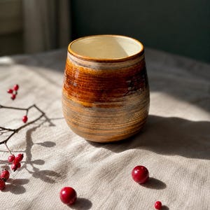 May include: A rounded ceramic tumbler featuring a glossy, multi-toned brown glaze with visible horizontal brushstroke textures and a smooth, cream-colored interior. The exterior surface displays a gradient of deep amber and earthy brown hues.